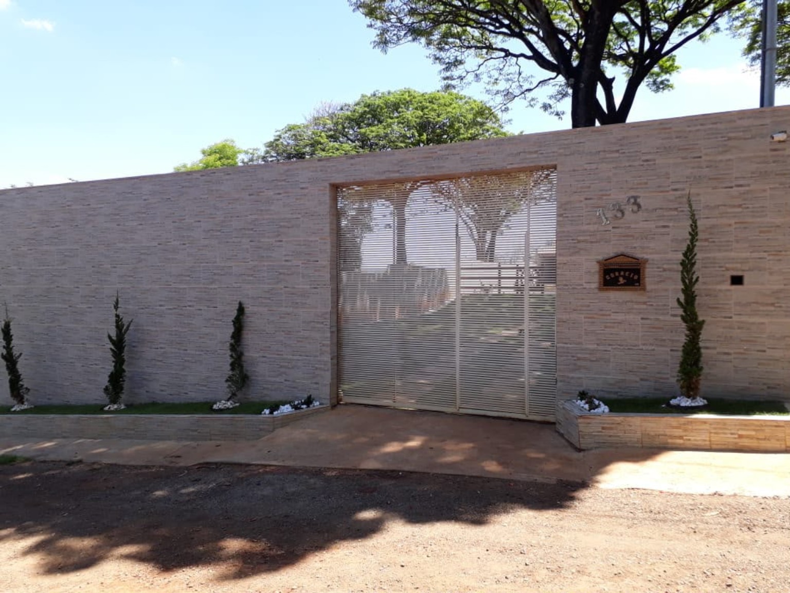Entrada do local da comemoração, com muro claro, portão metálico e o número 133